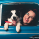 Una signora con occhiali, sorridente seduta all'interno di un'auto blu tiene in braccio un piccolo cagnolino bianco e nero, visibile attraverso il finestrino.